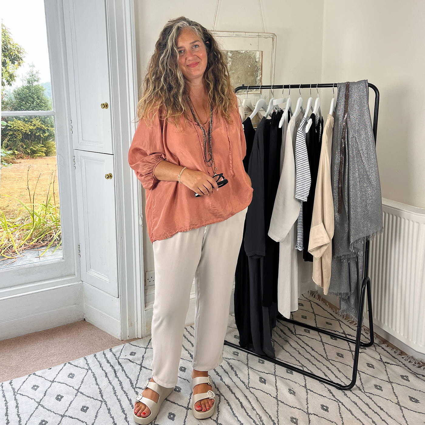 Emma wearing coral blouse and neutral trousers.