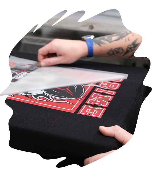 Hand peeling the clear film backing away from a black t-shirt revealing a bold red and white Prime logo DTF transfer