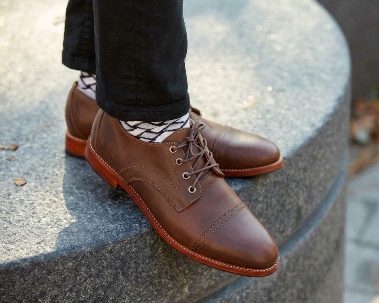 A close-up outdoor shot of a man's feet crossed at the ankles, resting on a curved grey stone structure. He is wearing brown or mahogany leather Adelante Marco cap-toe derby shoes with matching brown laces and a distinct leather sole. He is styled in black jeans and black-and-white patterned socks, with a paved walkway softly blurred in the background.