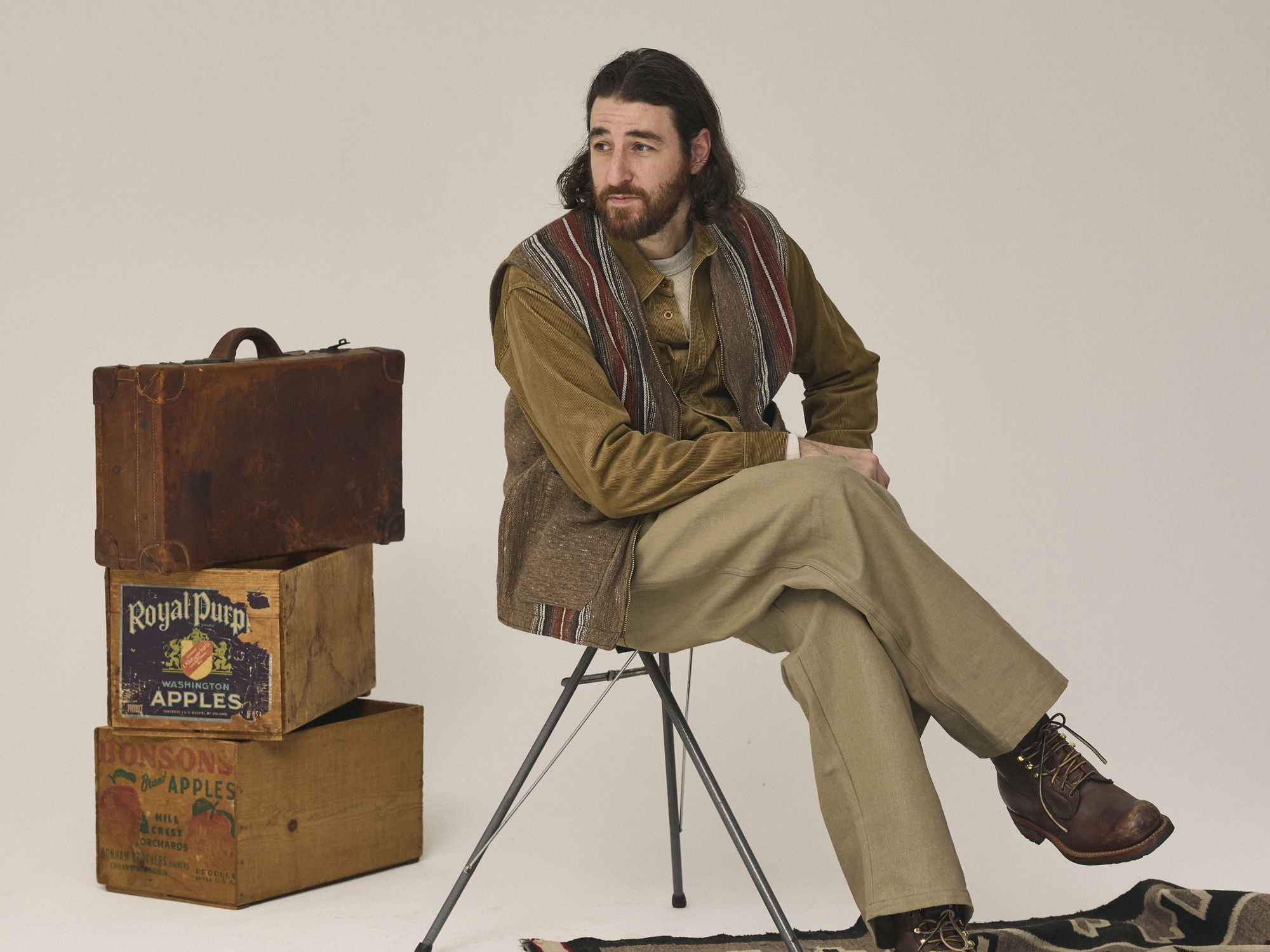 Man sitting on chair next to stacked wooden suitcases against plain background.