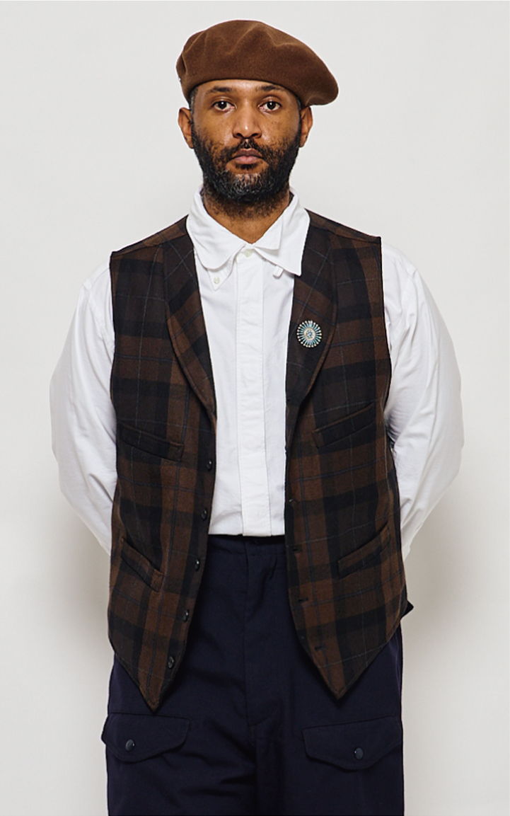 Man wearing a plaid vest, white shirt, and brown beret, standing against a neutral background.