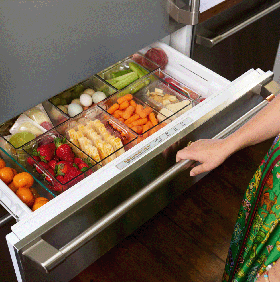 A hand reaching for a drawer full of fresh fruits and vegetables.