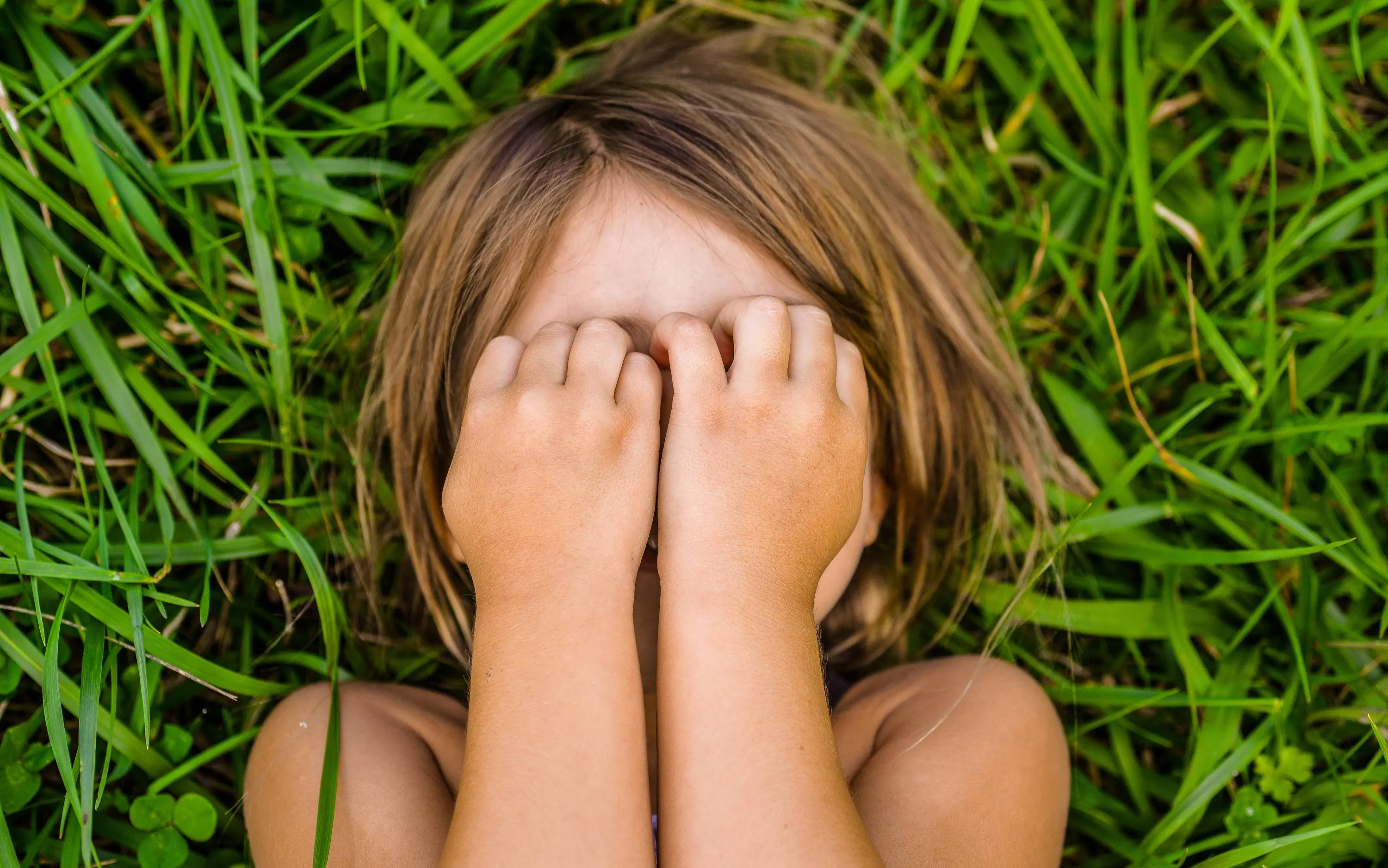Girl lying on lush green grass covering her eyes with her hands – it’s summer and grass pollen can trigger kids’ allergy eyes