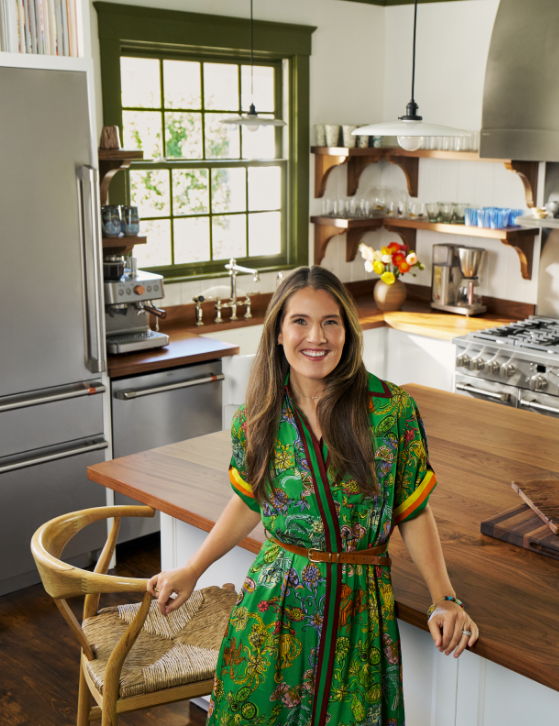 Smiling woman in a colorful dress stands in a bright kitchen.