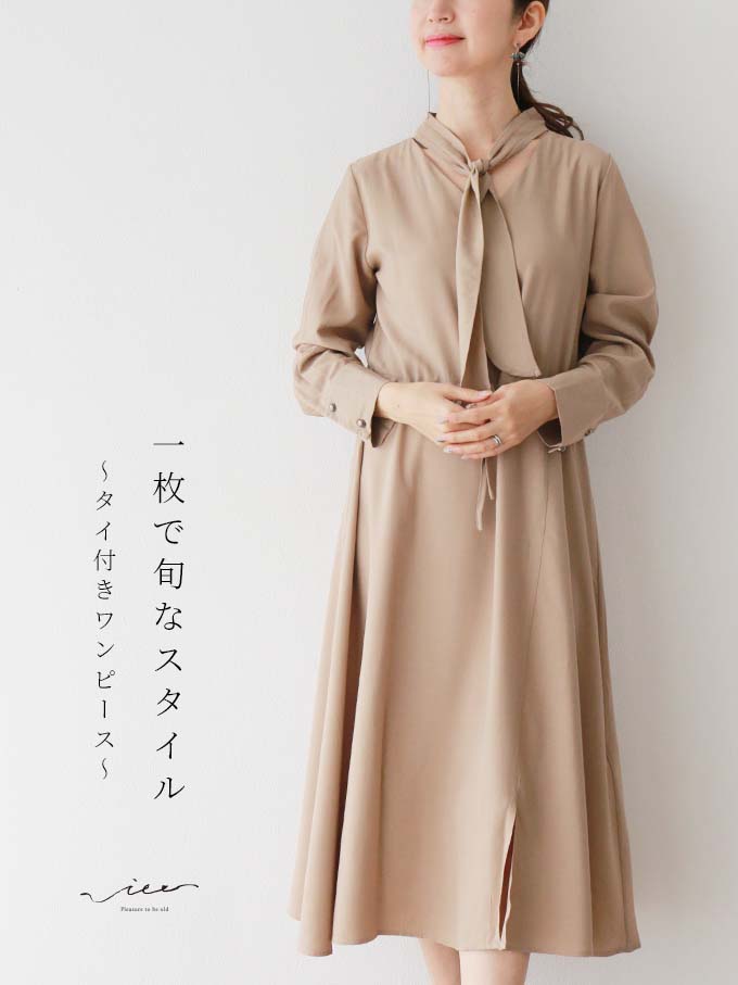 Woman in a beige dress with a tied collar, standing gracefully.