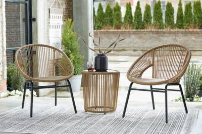 Two light brown coloured chairs that resemble wood or branches surround a small outdoor side table with a similar theme. On the table is a black vase with light brown branches emerging from the opening. The background is a concrete patio with shrubbery emerging from the wall.