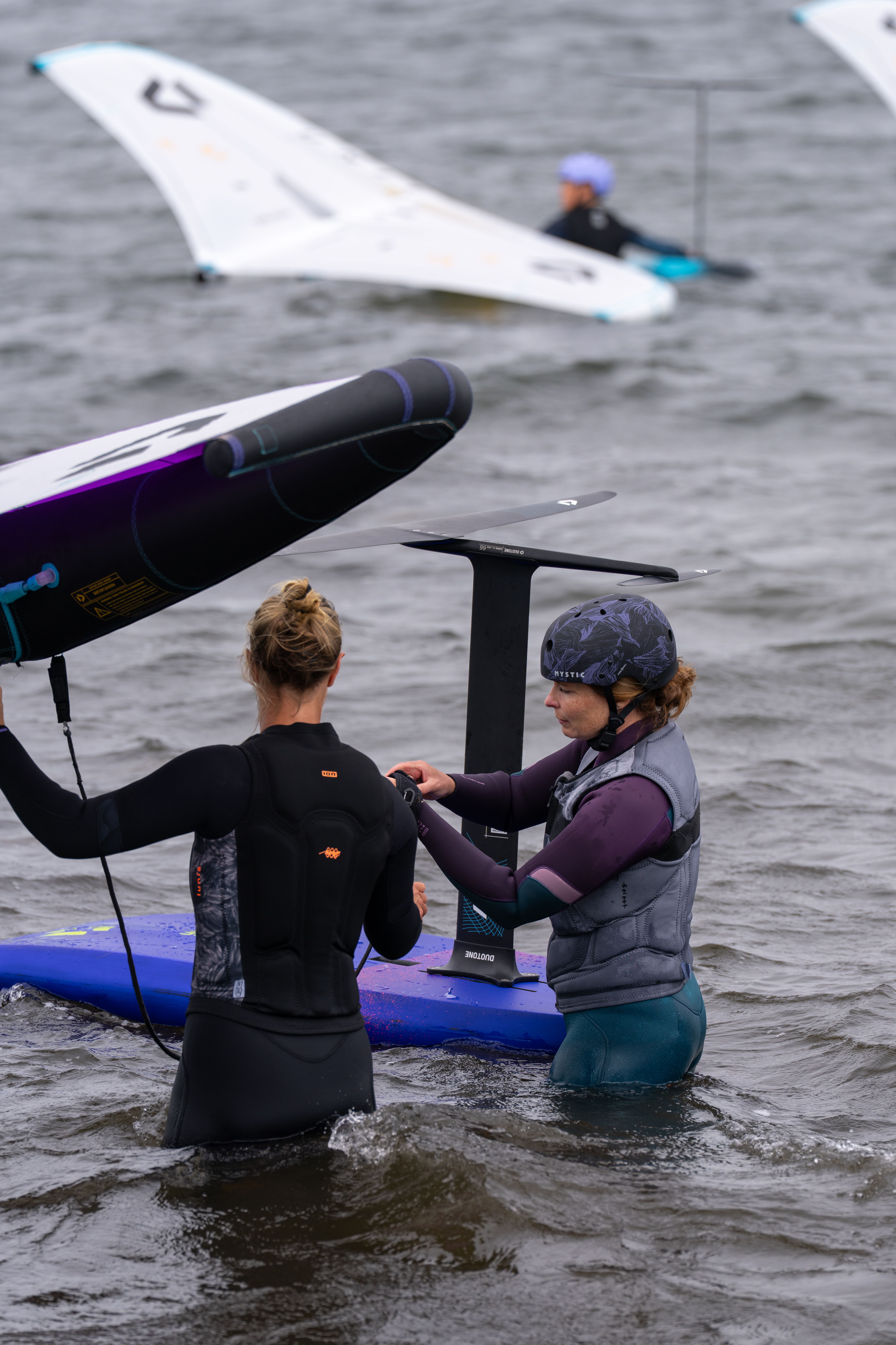 Two women preparing equipment while standing in water, with sails in the background.