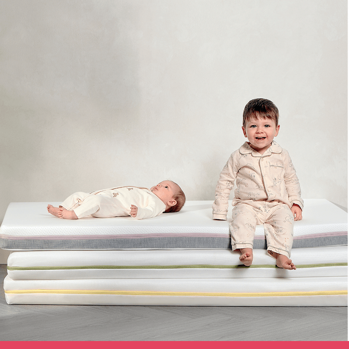 Two children on stacked mattresses, one sitting and smiling, one lying down.