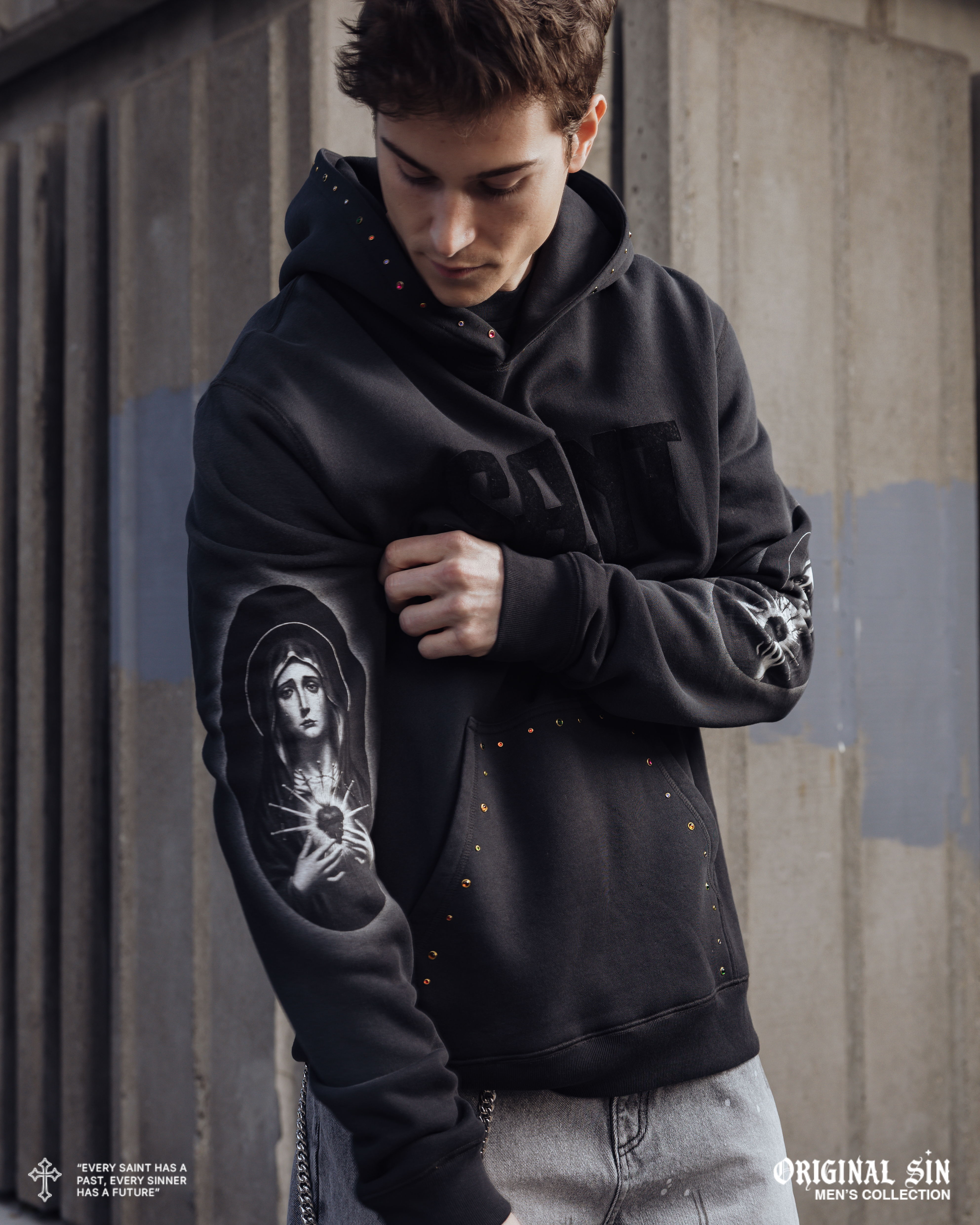 Young man in black hoodie with graphic design, looking thoughtfully at his arm.