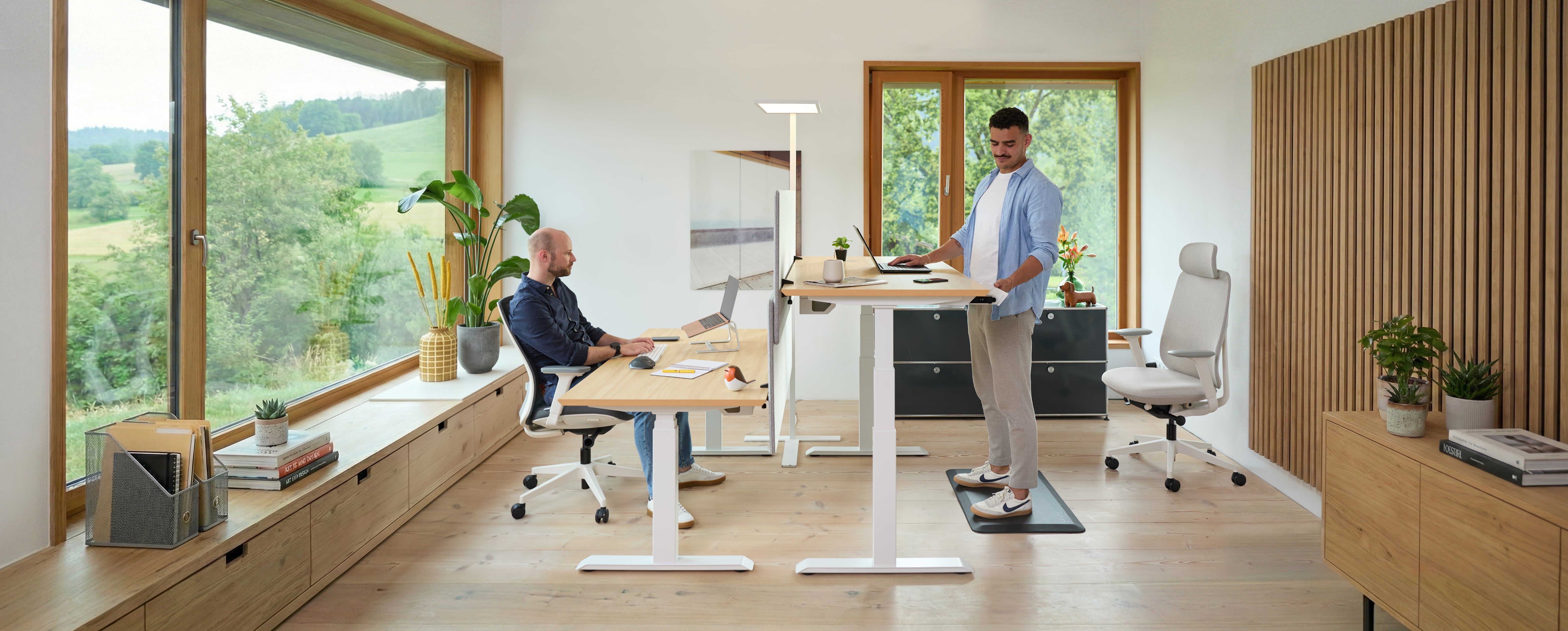 Two men working from home—one is sitting at a white ofinto Ergo, the other is standing on an ofinto standing mat. Both are working at ofinto Elevate desks Elevate a bright, modern room