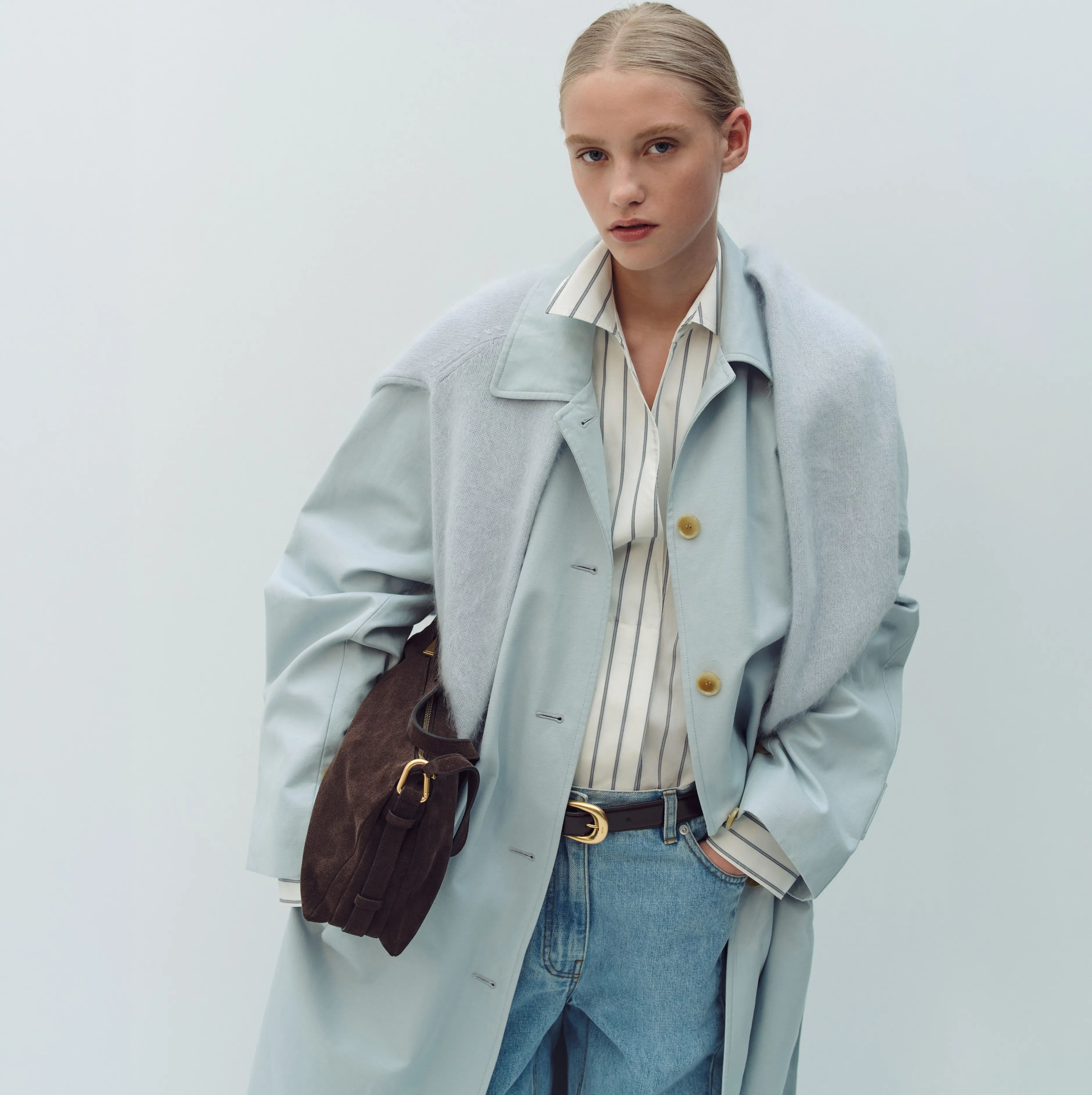 A woman in a light blue coat holds a brown handbag.