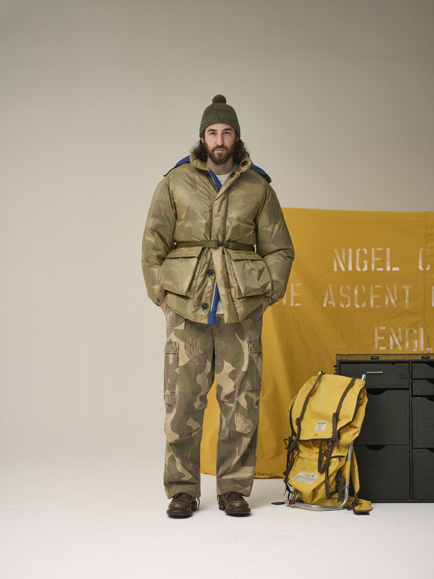 Person in winter attire standing next to a yellow banner and a backpack.