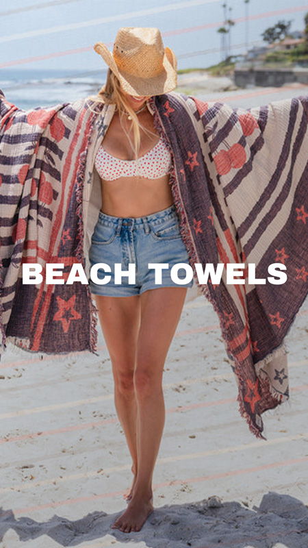 Person wearing a hat and beach attire, holding beach towels by the ocean.