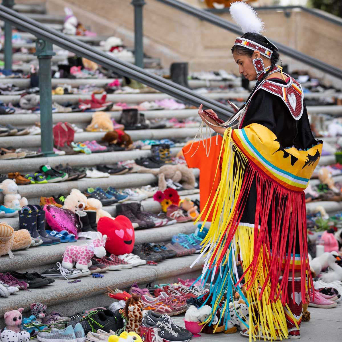 An Indigenous person in Traditional attire looking at steps covered in children's shoes