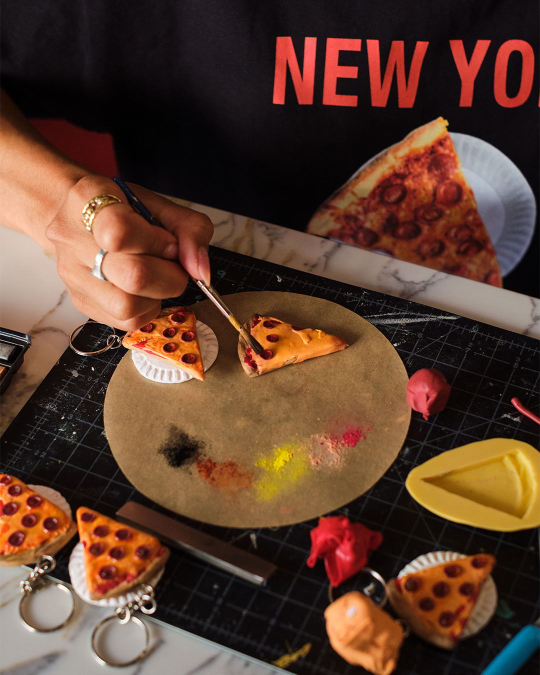 Person crafting miniature pizza on a workspace with various art supplies.