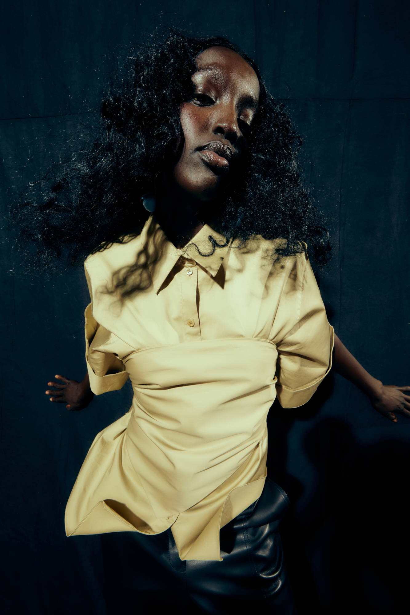 A person with curly hair wearing a yellow shirt against a dark background.
