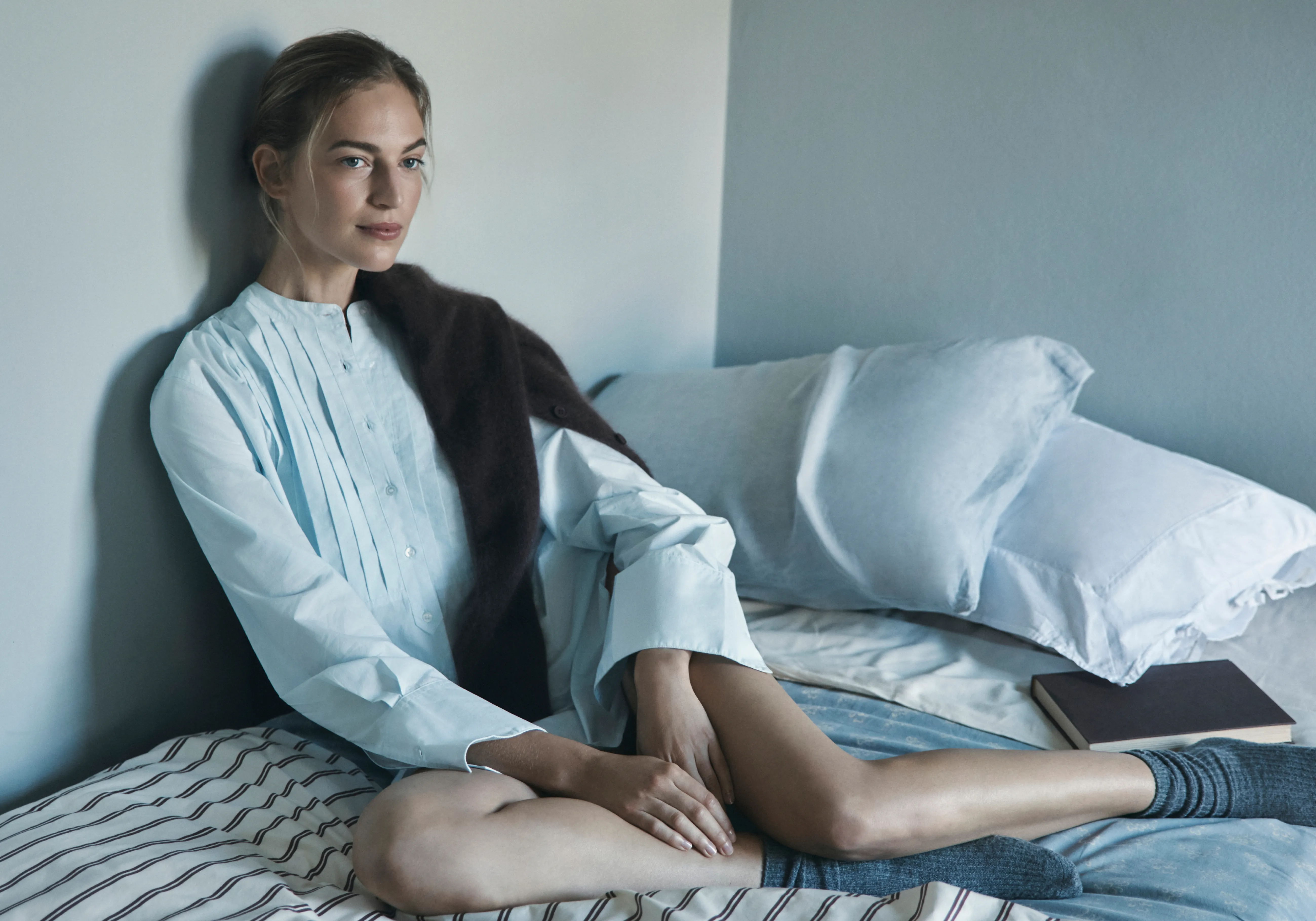A woman sitting on a bed, wearing a light blue shirt.