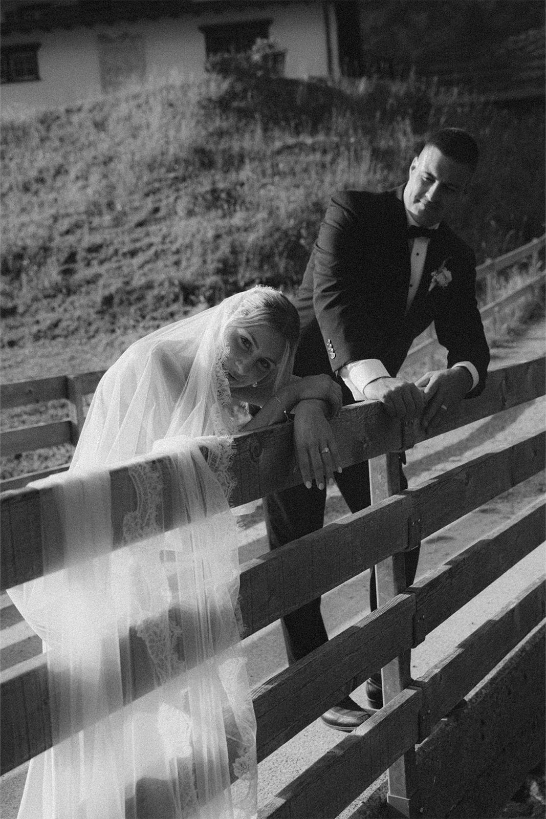 Bride and groom leaning against a wooden fence in black and white.