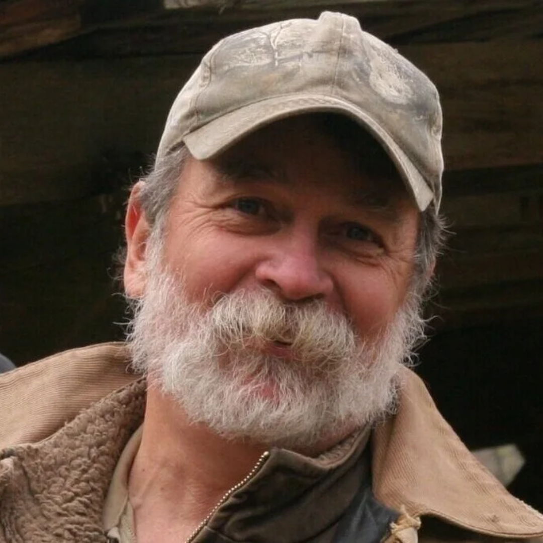 Smiling older man with a beard wearing a cap and casual clothing.