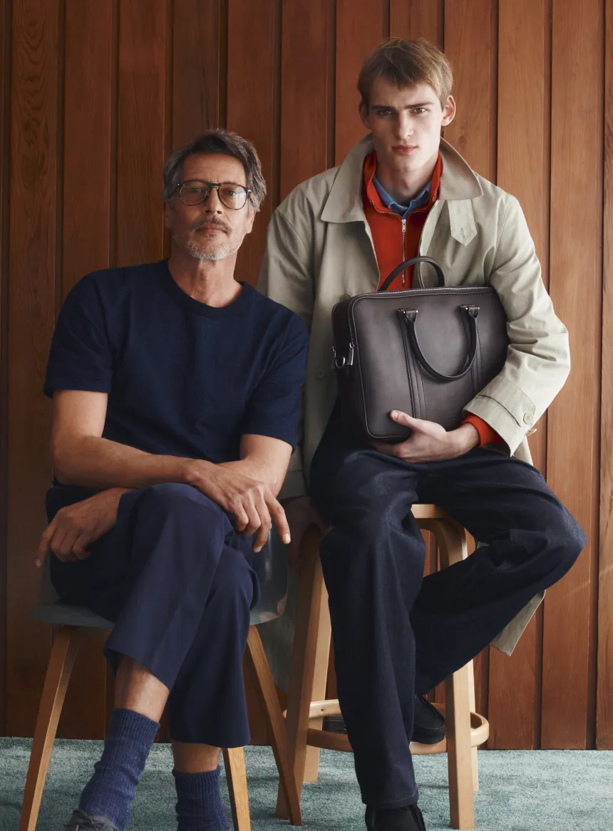 Two men sitting on stools against a wooden wall, one holding a bag.
