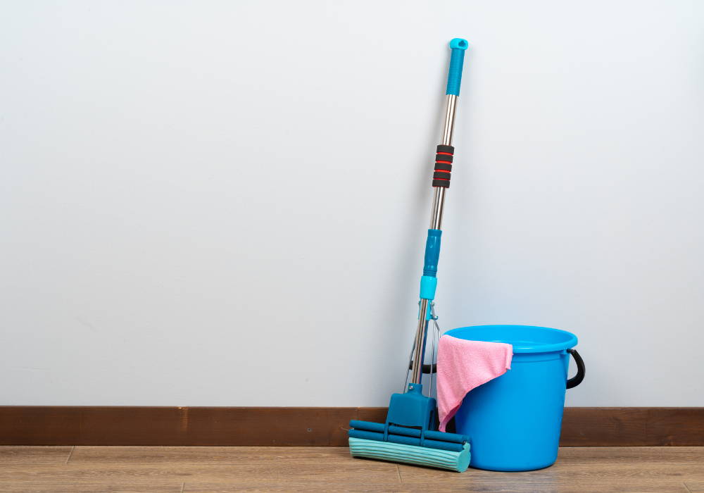 A blue sponge mop leaning against a wall and a blue bucket on a hardwood floor