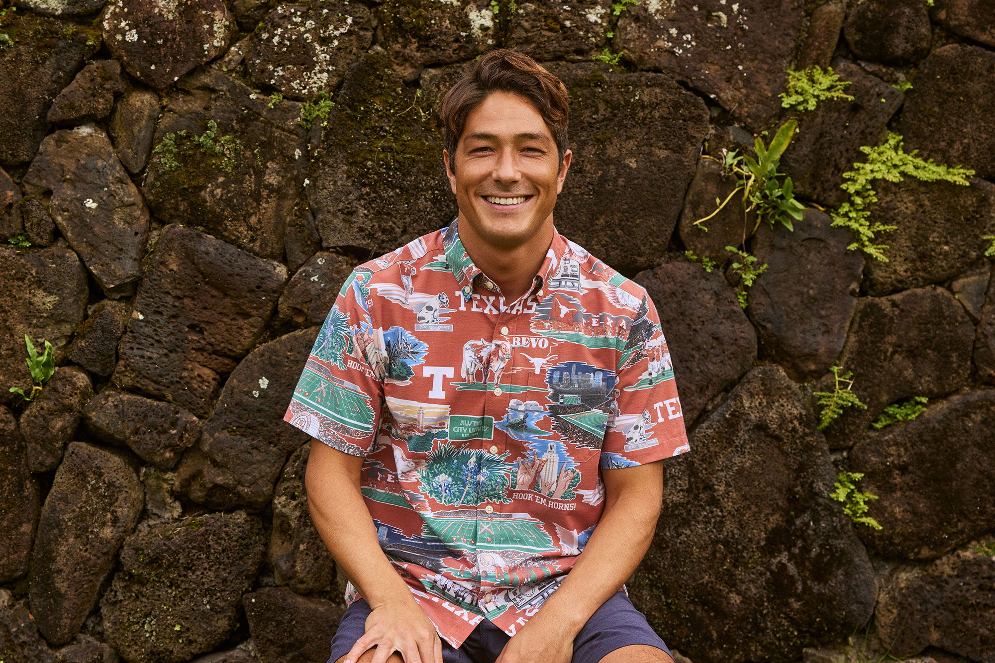 Smiling person in a colorful shirt sitting against a stone wall.