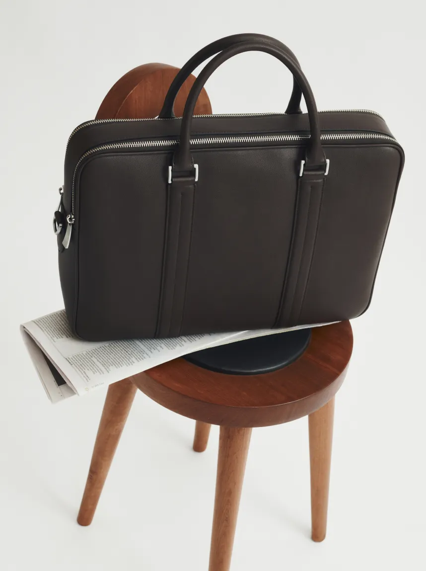 A brown leather bag resting on a wooden chair with a newspaper underneath.