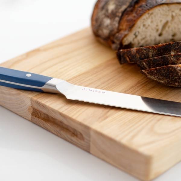 A blue Misen Serrated Knife resting on a light wood cutting board, with a sliced loaf of rustic bread visible in the background.