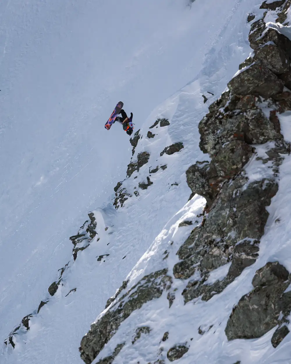 Snowboarder jumping off a rocky slope in a snowy mountain landscape.