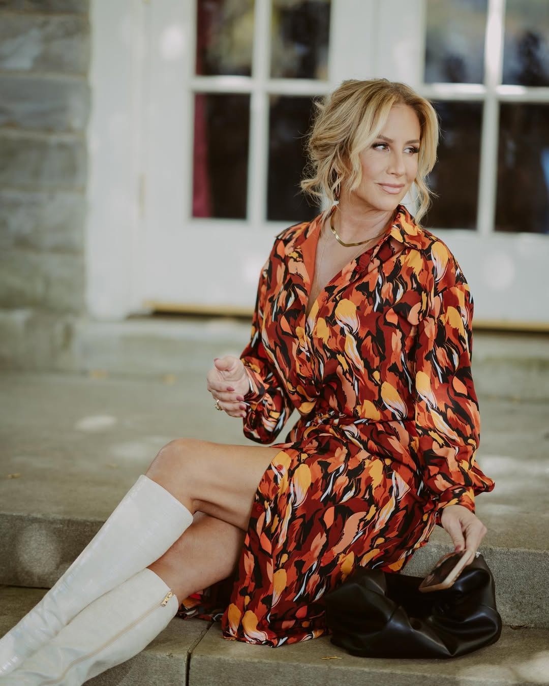 Woman in colorful dress sitting on steps, wearing white boots and holding a bag.