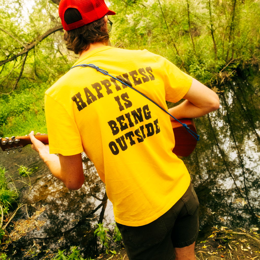 A person in a yellow shirt playing guitar by a pond.