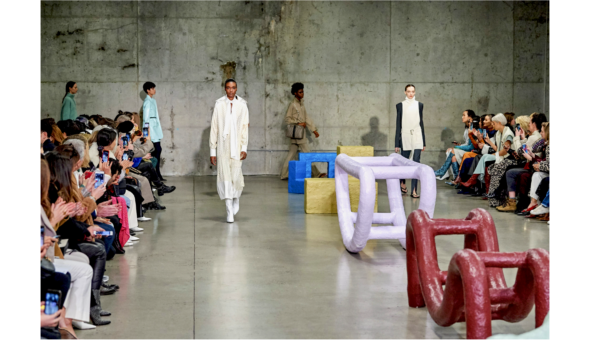 Fashion show with models walking past an audience in a concrete setting.