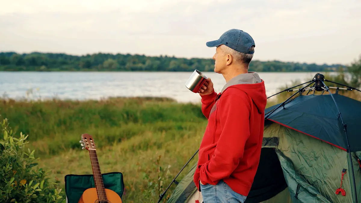 Person in a red jacket enjoying a drink by a lakeside campsite.