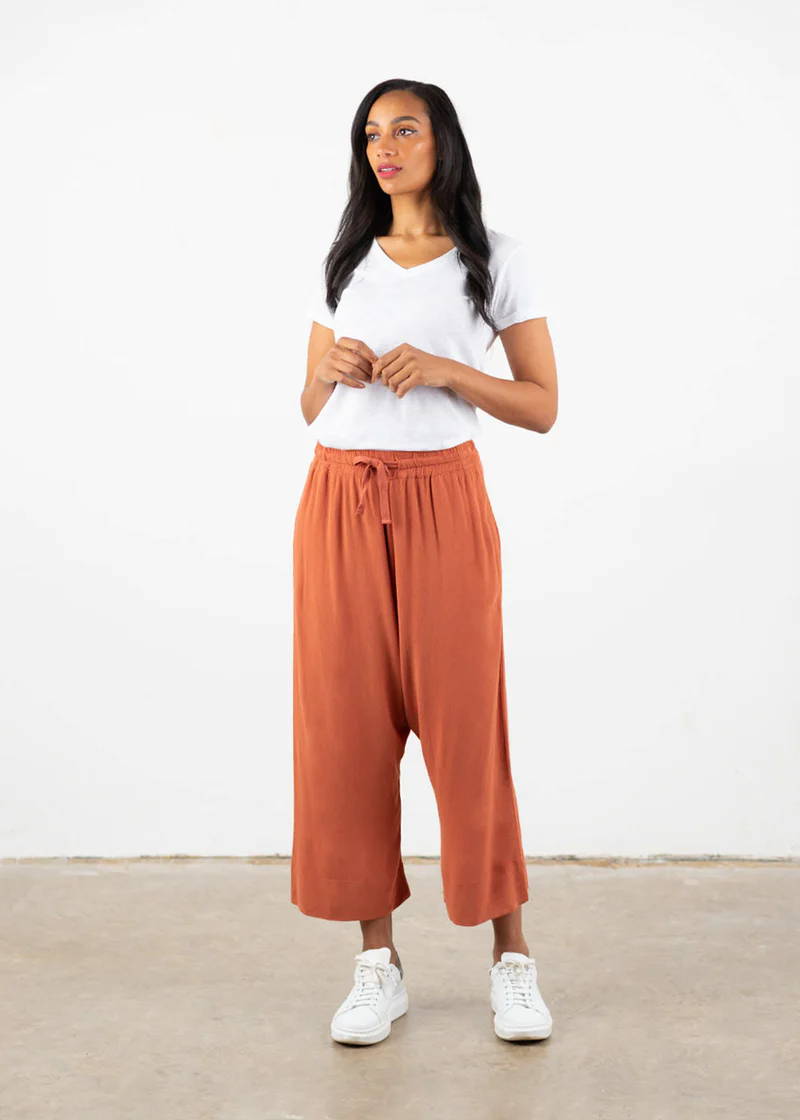A model wearing a pair of terracotta loose fit cropped trousers with a white t-shirt and white trainers
