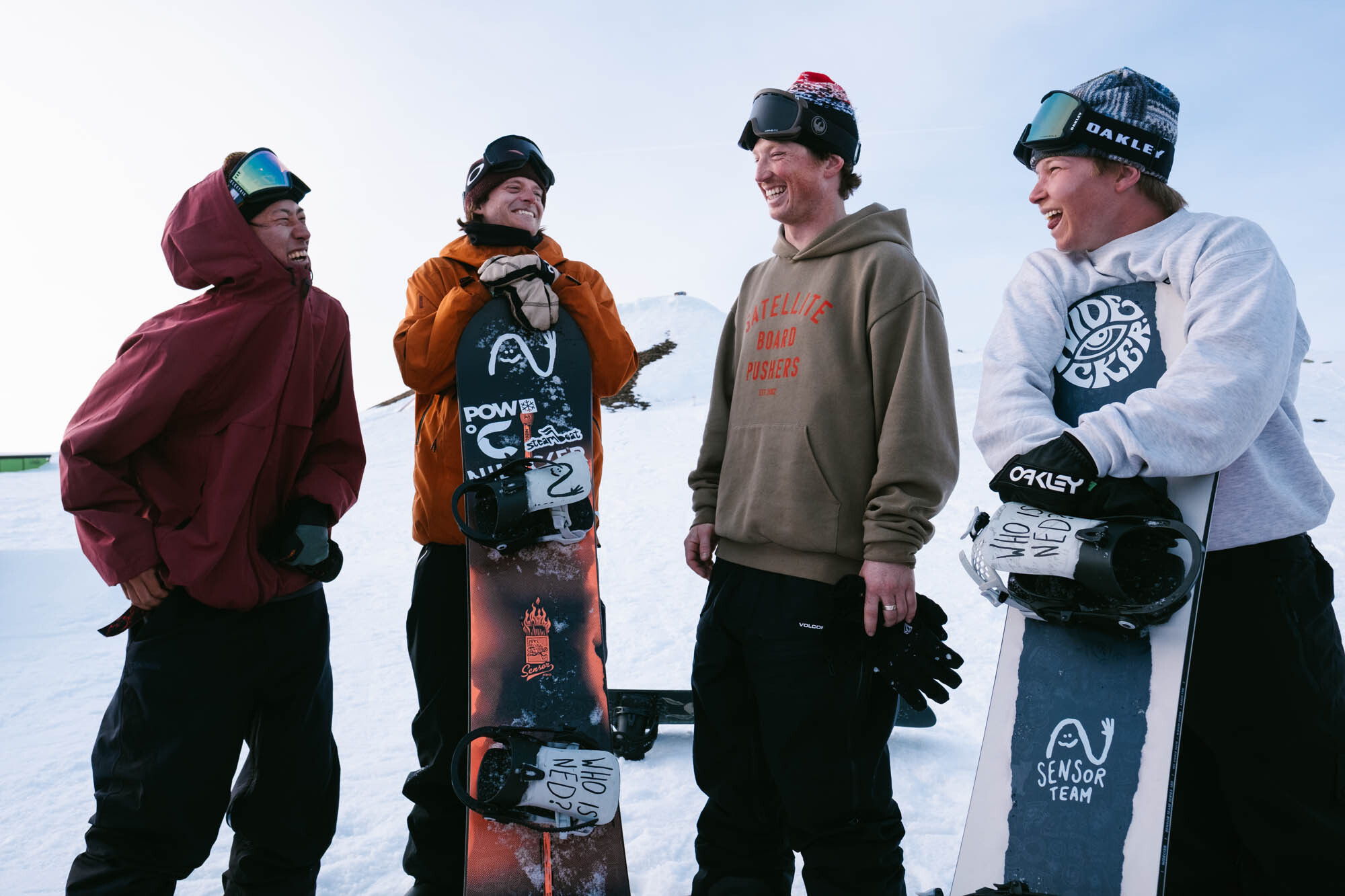 Four snowboarders chatting on a snowy slope, wearing winter gear and helmets.