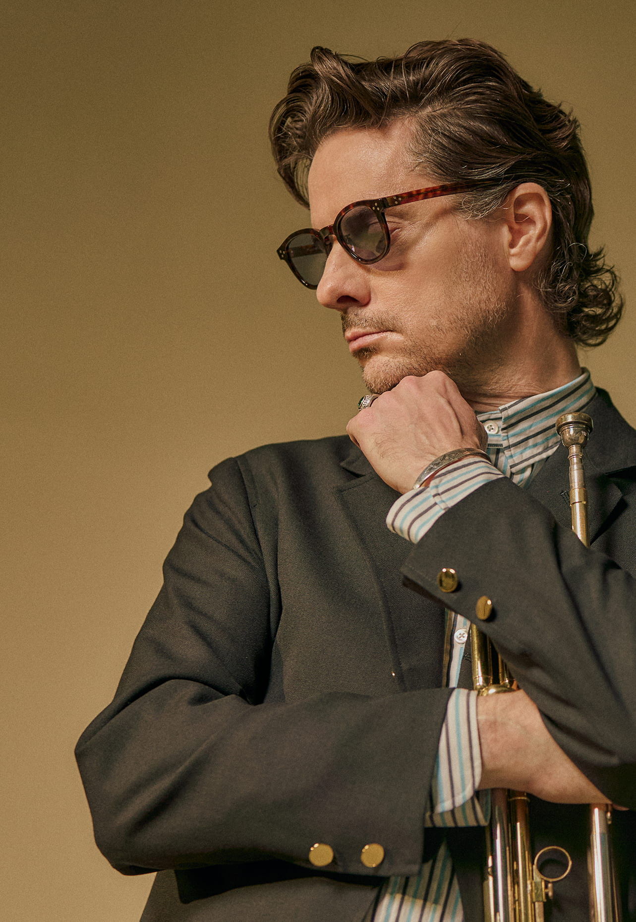 Man in sunglasses with a trumpet, striking a thoughtful pose against a plain background.