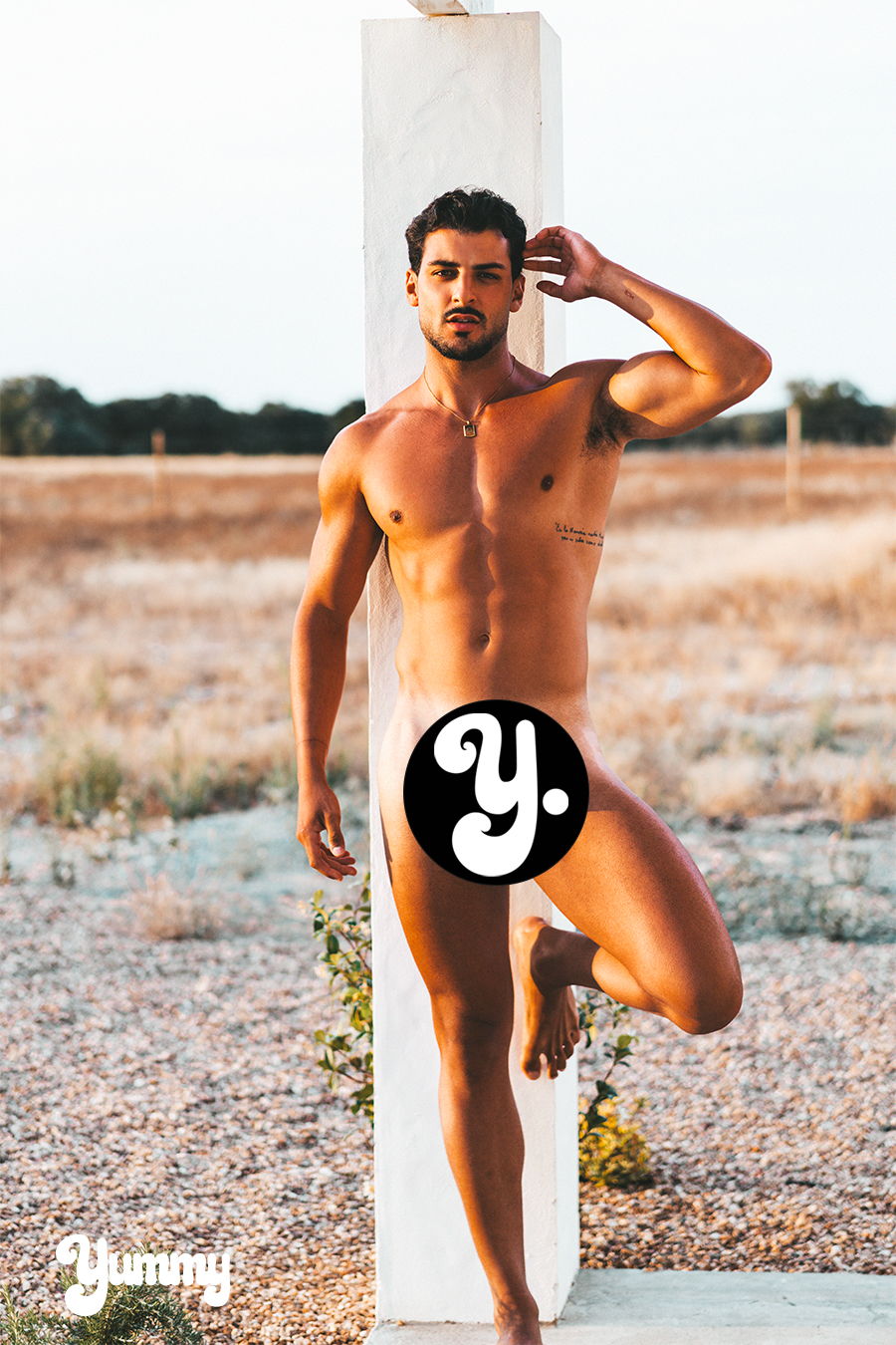 A man posing nude beside a vertical structure in an outdoor setting.