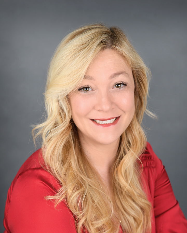 Smiling woman with long blonde hair wearing a red blouse against a gray background.