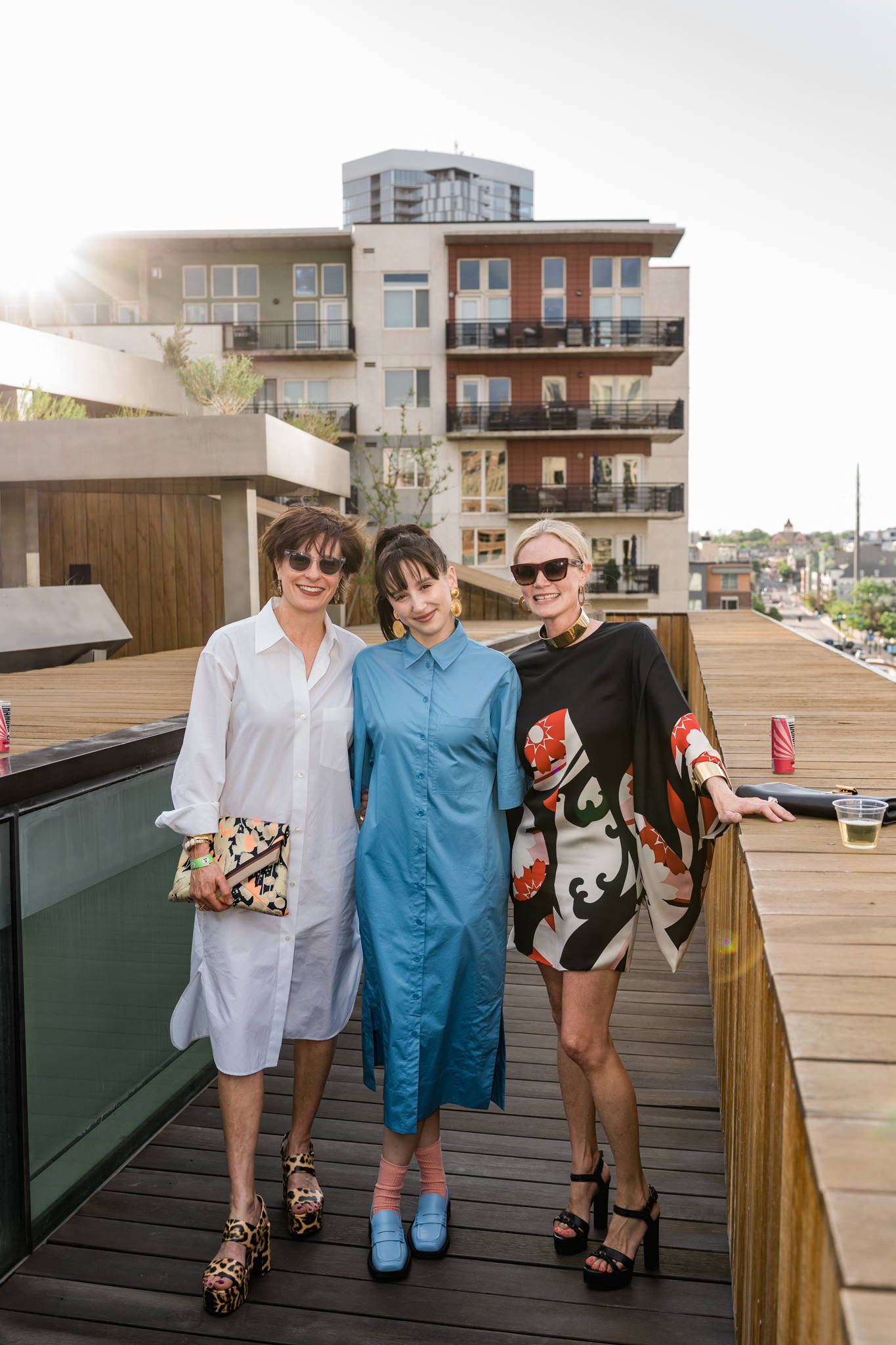Three women in stylish outfits pose on a rooftop terrace.