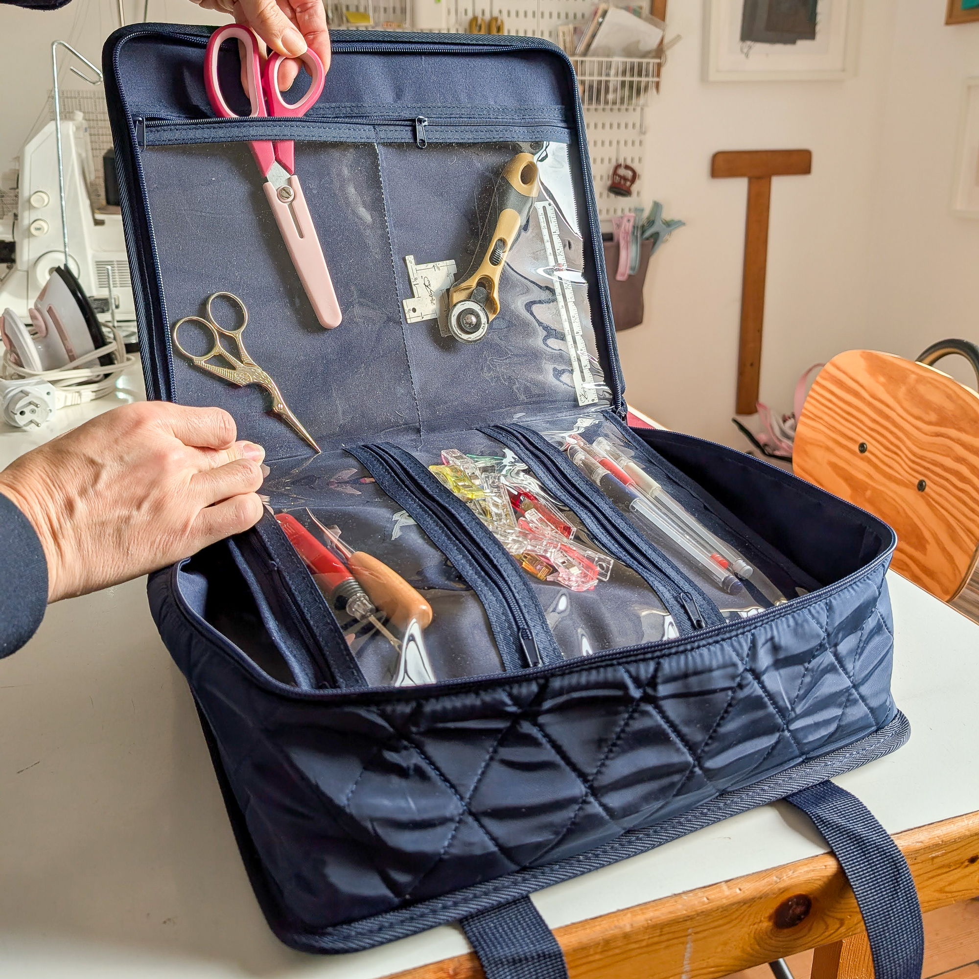 a blue carry-all sewing organizer on a sewing table holding sewing tools in clear pockets with zippers