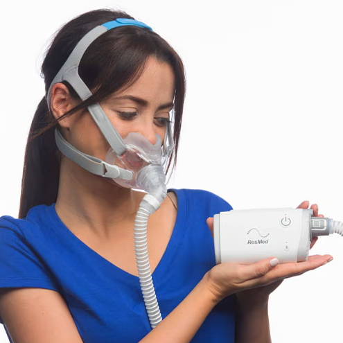 Woman in blue shirt holding a small CPAP and wearing a mask.