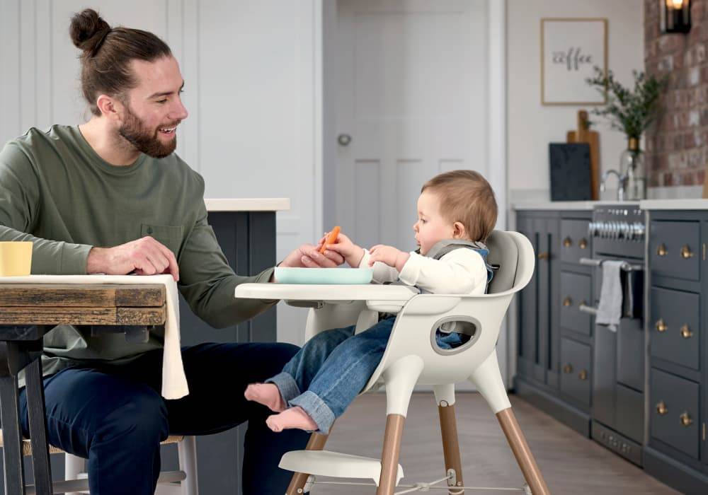 A dad sits with his baby who is in their Mamas and Papas Juice highchair.