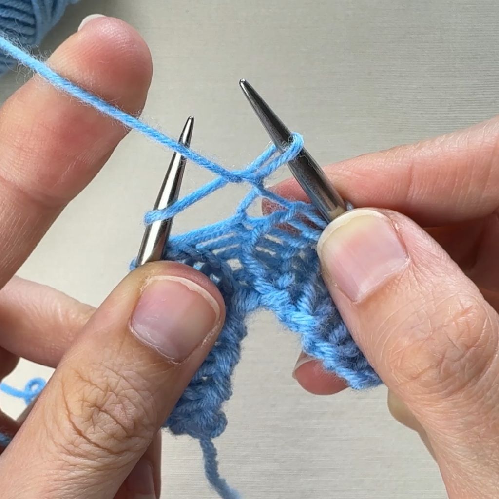 Detailfoto, wie eine linke Masche gestrickt wird. Die Masche liegt nun auf der rechten Nadel.