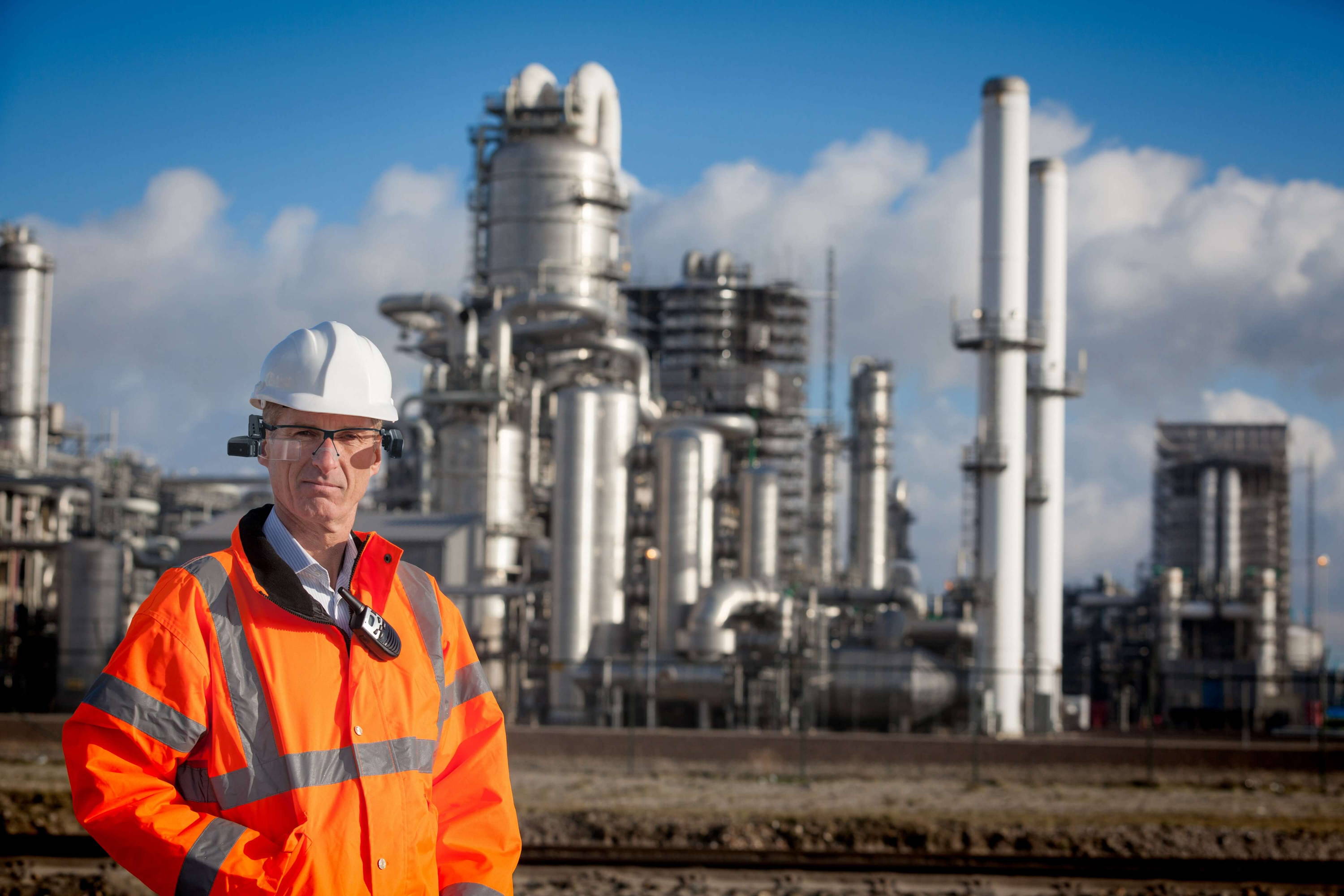 Man in orange safety jacket, white hard hat, and Vuzix smart glasses standing in front of a processing plant.