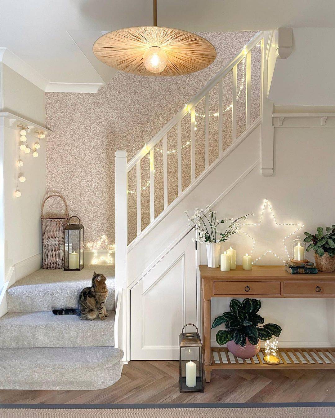 A staircase with fairy lights and star lights draped over the banister and a cat admiring them.