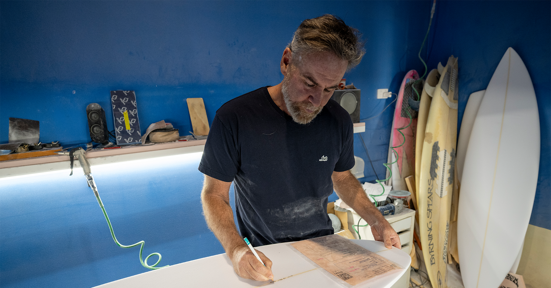 Person shaping a surfboard in a workshop with surfboards in the background.