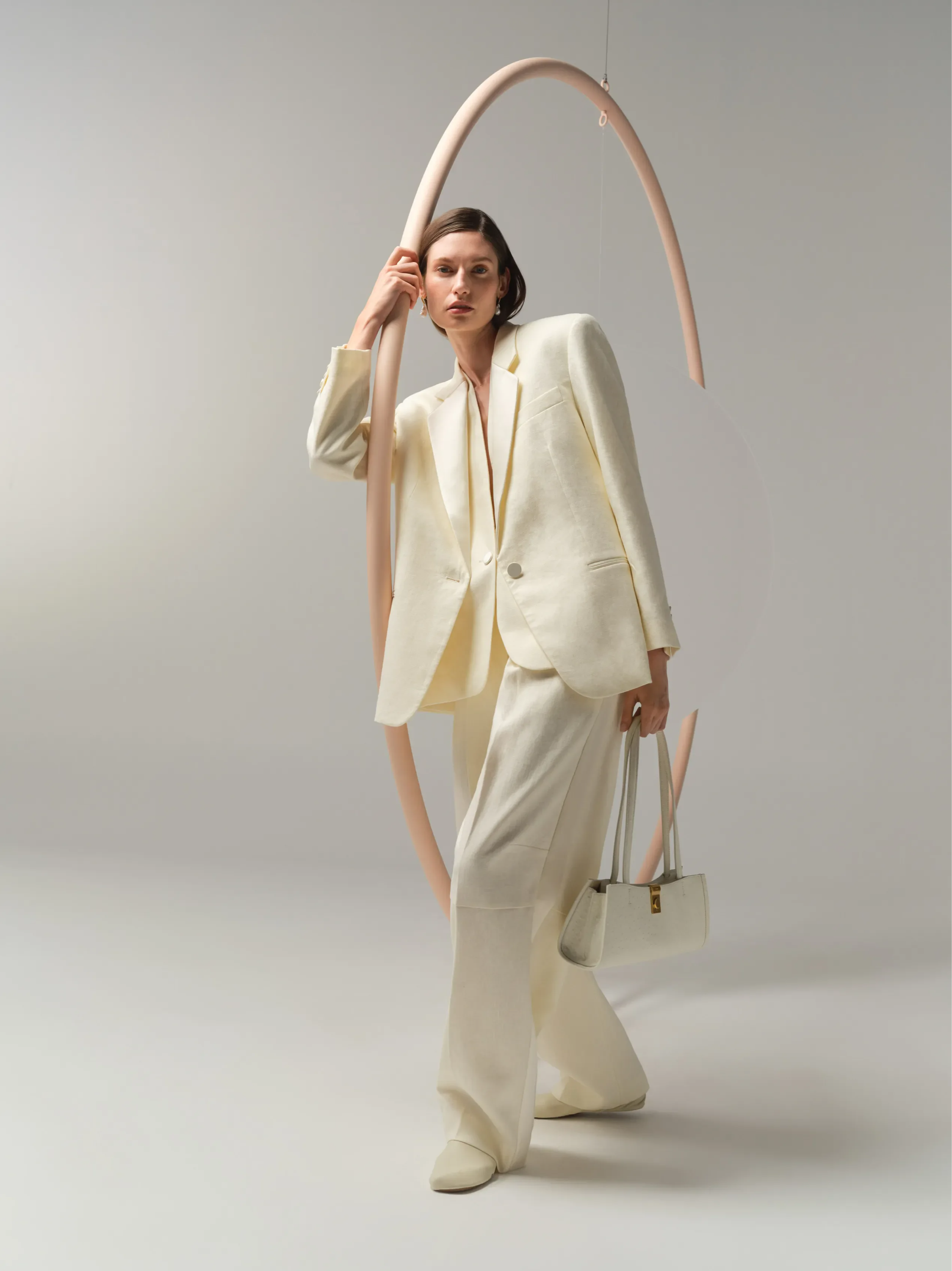 Model in a cream suit holding a handbag, posed with abstract shapes.