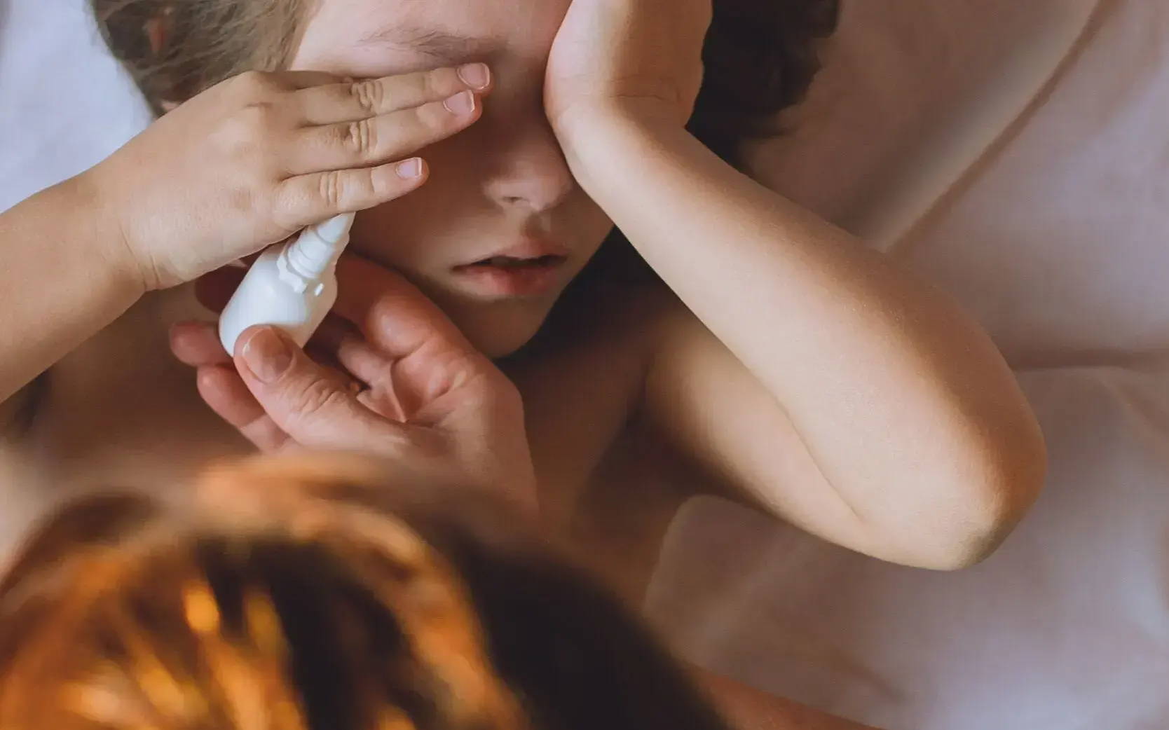  Dívka leží v posteli s rukama na obličeji, máma se nad ni naklání a dává jí oční kapky ke zklidnění svědění kvůli alergii