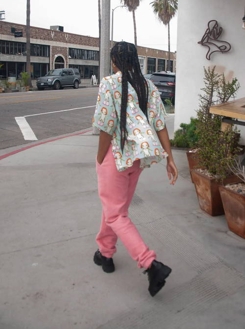 Photo of woman with braids walking away from camera, wearing an anime print Sandy Liang button up shirt and pink Brain Dead sweatpants.