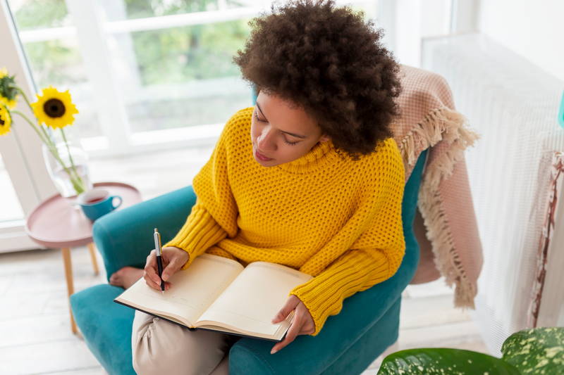 Vrouw met kroeshaar en gele trui maakt zittend in een fauteuil een schema dat ze in een planner opschrijft.